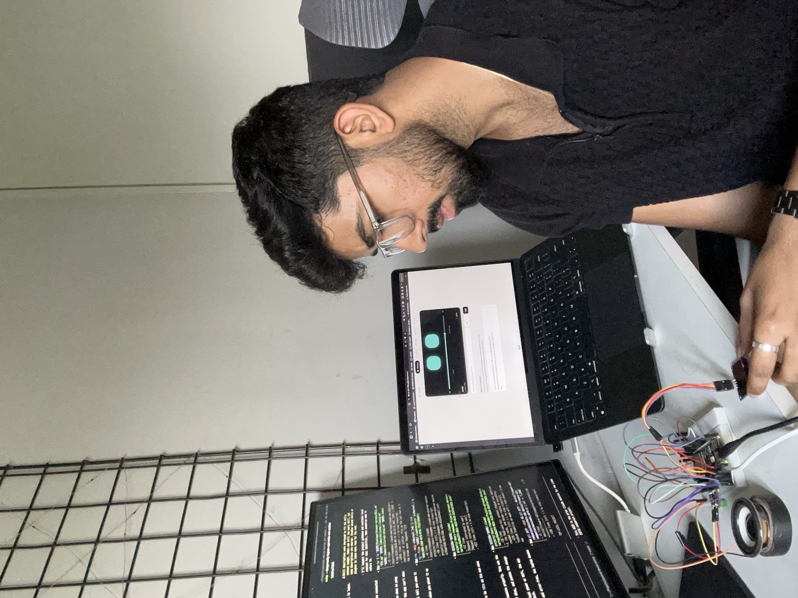 Working at a desk with a laptop showing the companion's web app with two cyan eyes on screen, and the ESP32-S3 prototype on a breadboard in the foreground with jumper wires, a speaker, and an OLED display